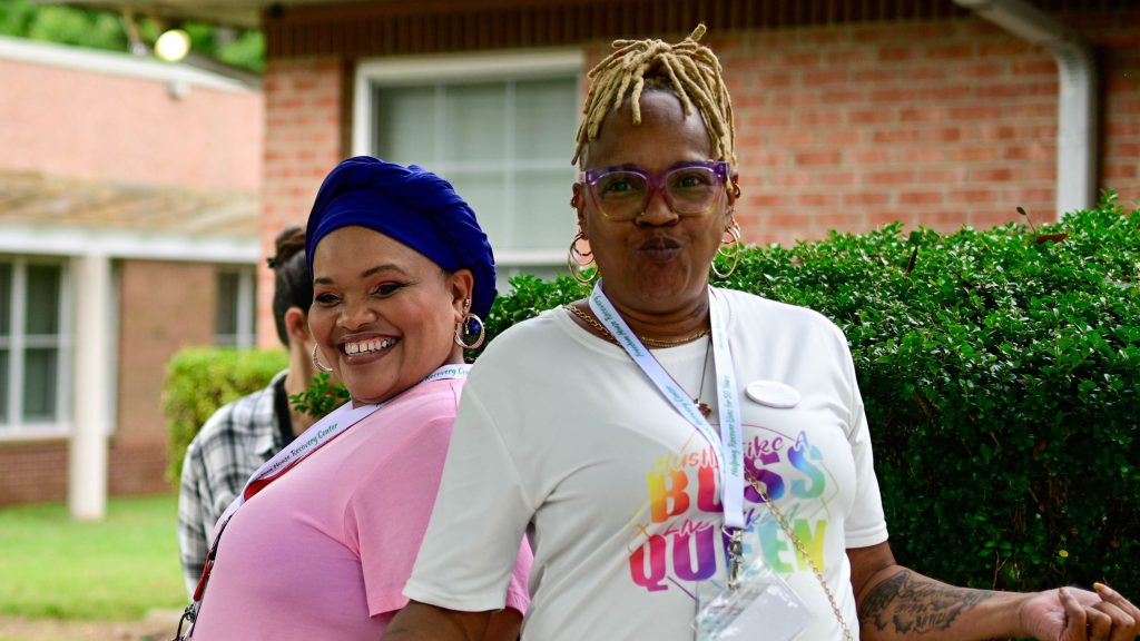 Two smiling women standing back to back outside near bushes and a brick building
