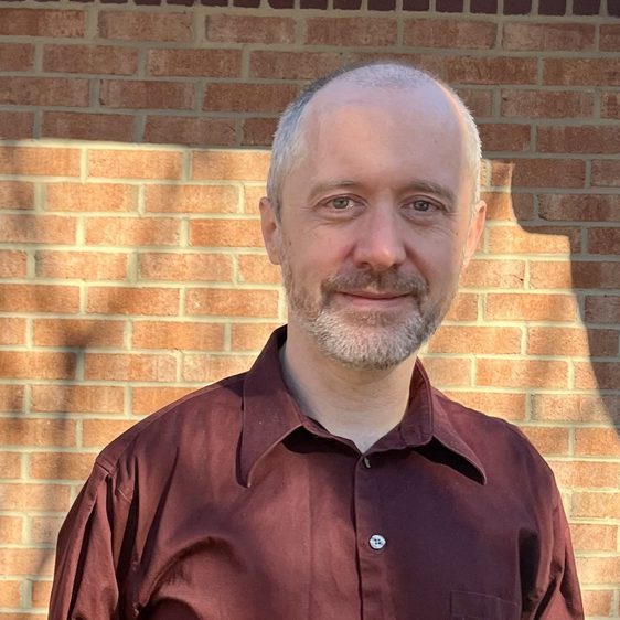 Smiling man with short beard and maroon shirt standing against brick wall