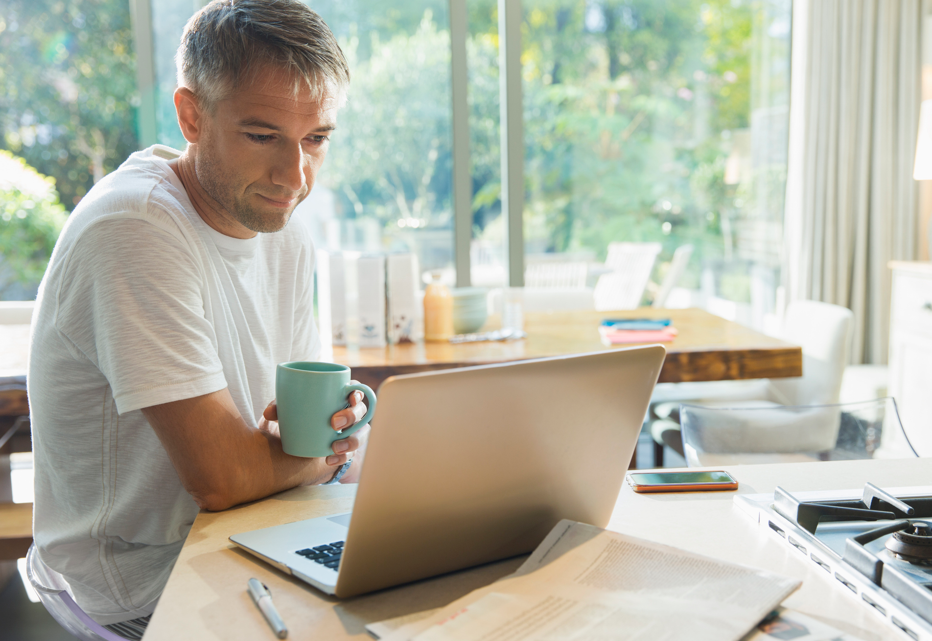 Man in white shirt holding mug and looking at laptop at kitchen counter