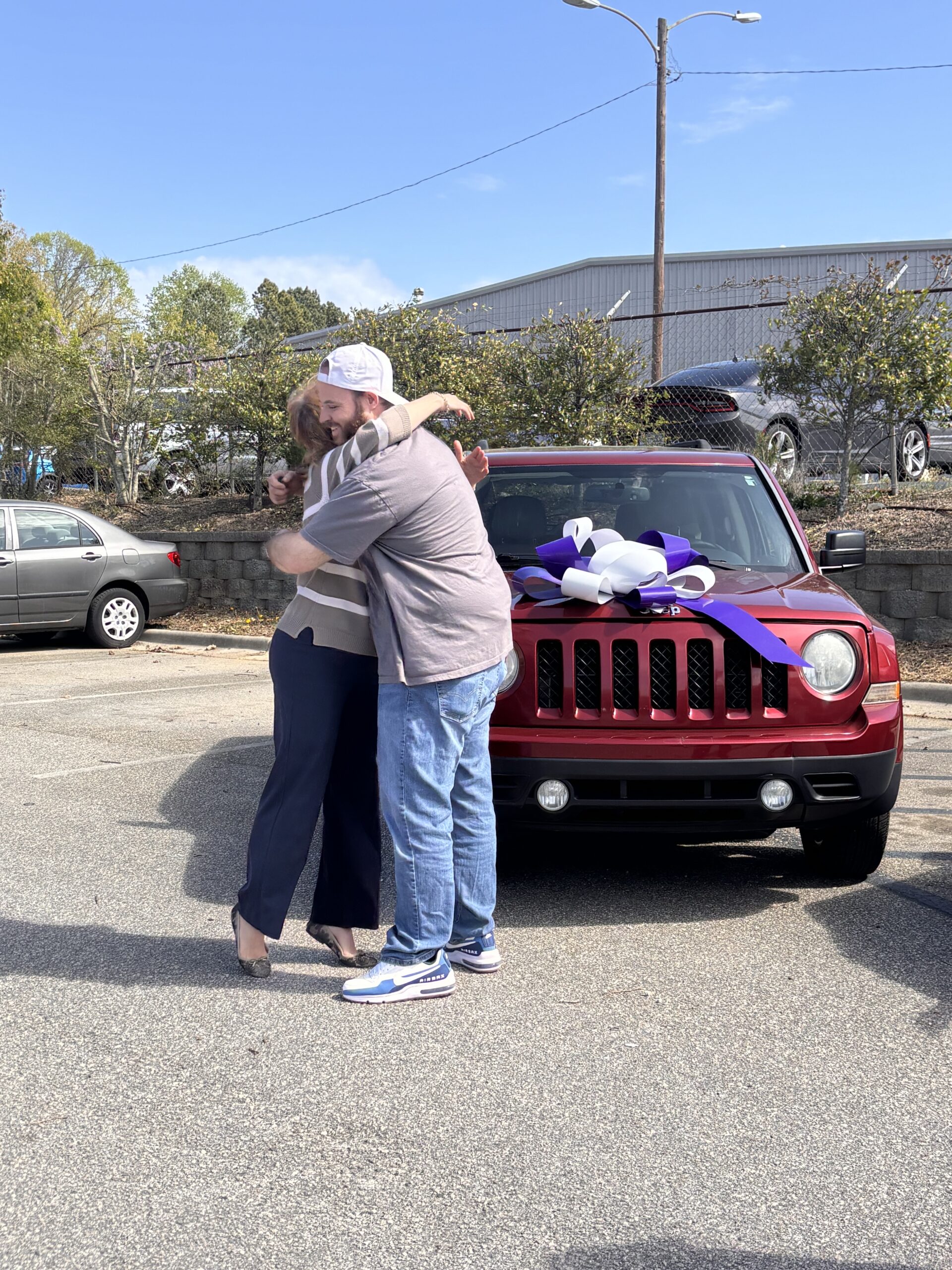 Man and woman hugging in front of red Jeep with large purple and white bow