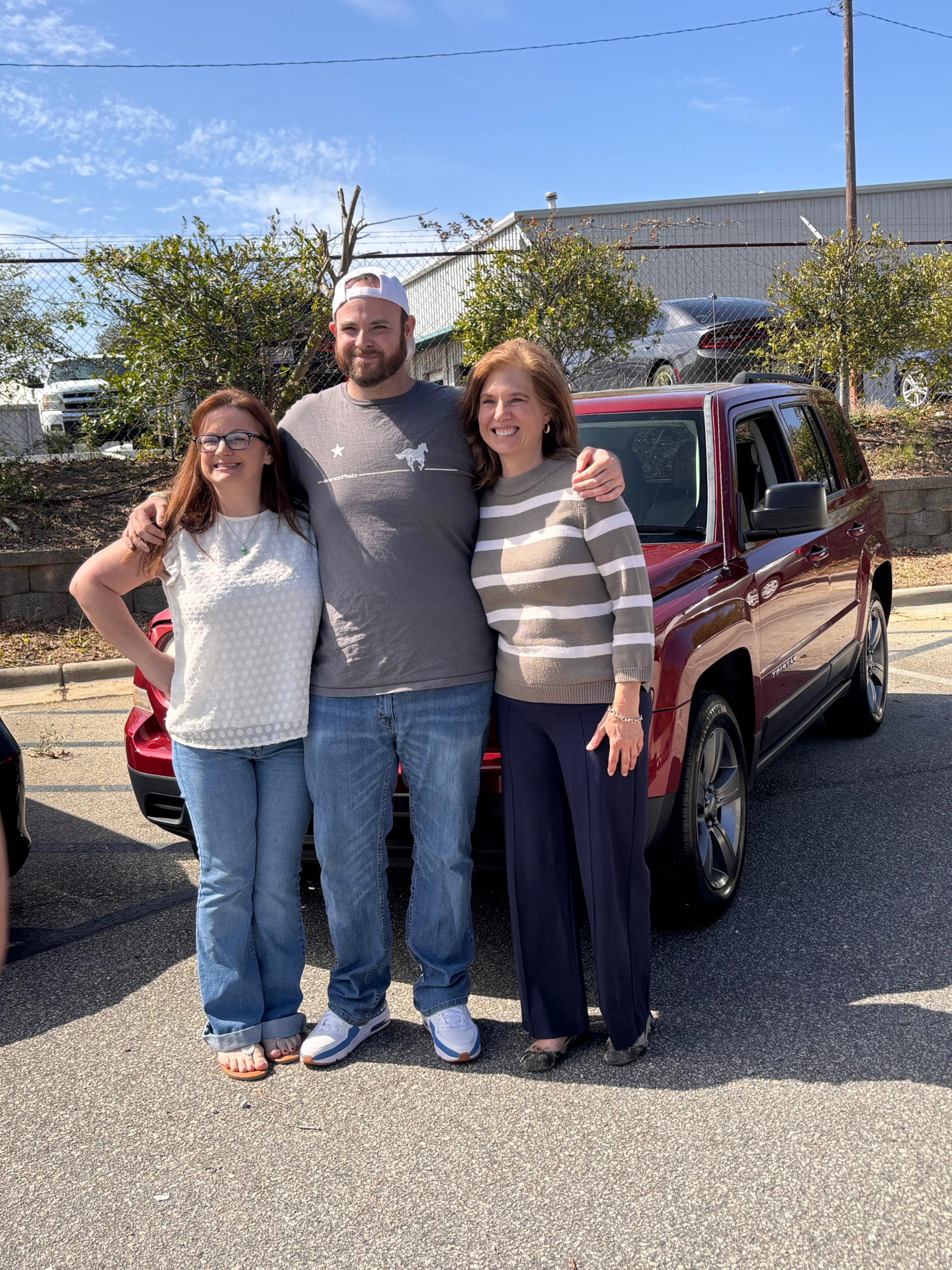 Three people standing together and smiling in front of a red SUV on a sunny day