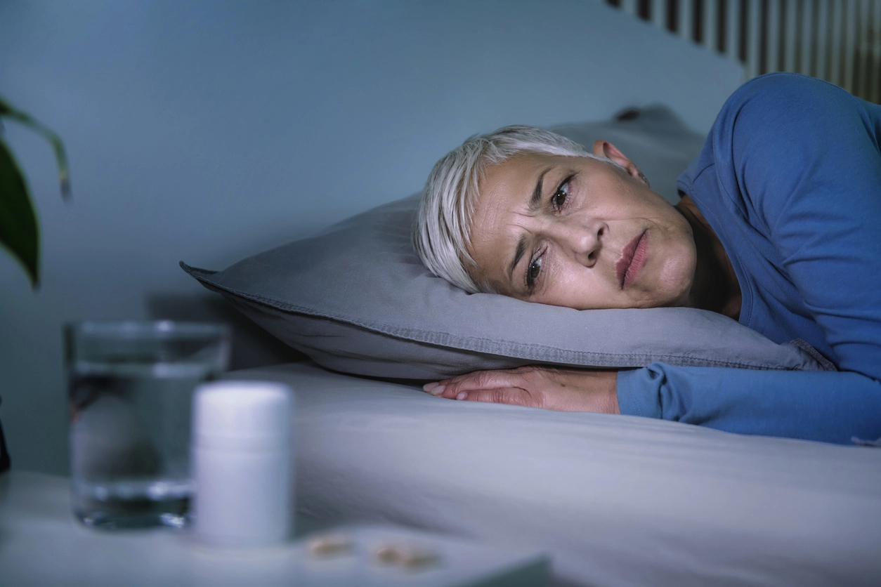 Woman lying in bed with a concerned look, staring at a water glass while her head is on the pillow.