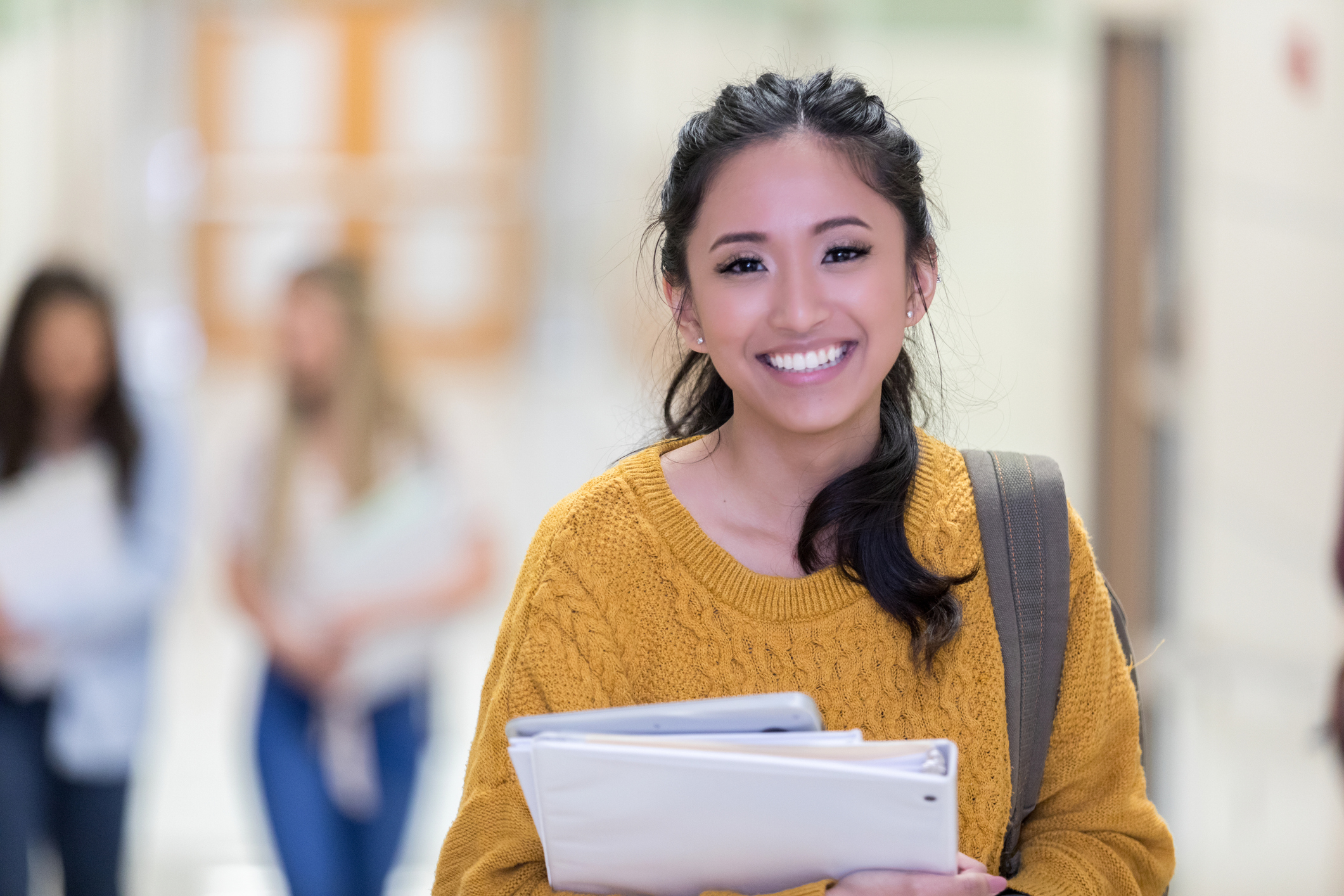 Young, smiling female student walks down hallway carrying books and a tablet.